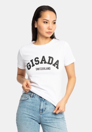 Femme portant un t-shirt blanc avec le texte "GISADA SWITZERLAND" et un jean bleu clair, debout devant un fond uni clair.