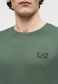 Κοντινό πλάνο ενός ατόμου που φοράει πράσινη μπλούζα με στρογγυλή λαιμόκοψη και μικρό μαύρο λογότυπο EA7 Emporio Armani στο στήθος.