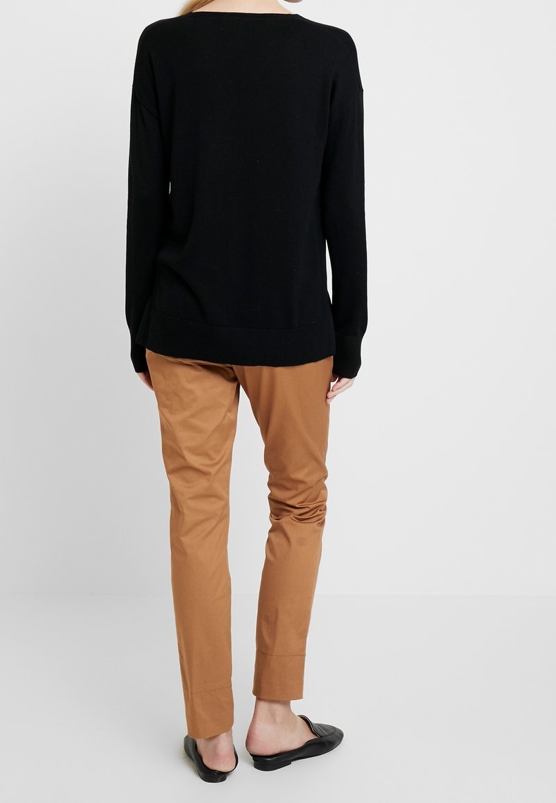 Personne debout de dos portant un haut à manches longues noir, un pantalon beige et des chaussures noires sans lacets sur un fond clair uni.