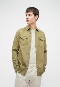 Olívazöld, gombos ing két mellzsebbel, világos bézs textúrált felsővel és krémszínű, megkötős nadrággal párosítva.