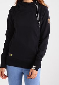 Kvinna iklädd en svart huvtröja med hög krage och dragsko, i kombination med ljusblå jeans, stående mot en enfärgad bakgrund.