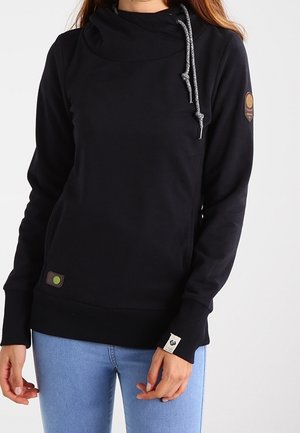 Frau, die einen schwarzen Hoodie mit hohem Kragen und Kordelzug trägt, kombiniert mit hellblauen Jeans, vor einem einfarbigen Hintergrund stehend.