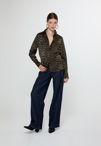 Leopard-print blouse in olive green with a collar, long sleeves, and a relaxed fit, paired with high-waisted dark denim wide-leg pants.