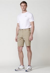 Pantaloni scurți bej ușori, cu buzunare frontale și închidere cu buton. Asortați cu un tricou alb ce are un mic logo negru. Adidași albaștri și gri.