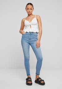 Valkoinen ribattupinta crop-top, jossa on avainaukko-yksityiskohta ja röyhelöhelma; vaaleansiniset korkeavyötäröiset skinny-farkut; mustat liukuva sandaalit paksuilla hihnoilla.
