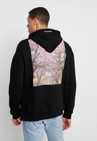 Black hooded sweatshirt featuring a large back print of pink cherry blossoms against a textured background. Ribbed cuffs and hem.
