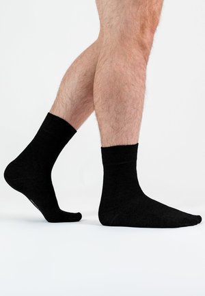 Männliche Beine tragen schlichte schwarze Crew-Socken, ein Fuß flach auf dem Boden, der andere leicht auf den Zehen erhoben, vor weißem Hintergrund.