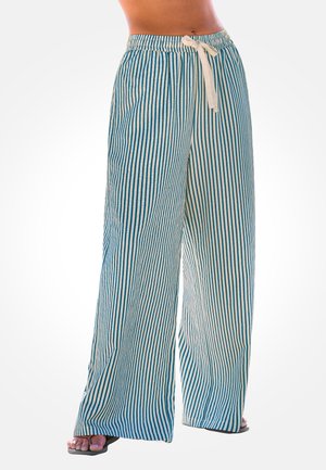 Woman wearing loose, wide-leg pants with vertical blue and white stripes and a white drawstring, standing barefoot on a white background.