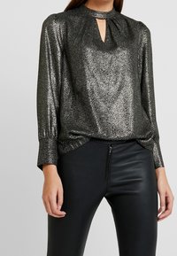 Blusa nera lucida realizzata in tessuto metallico, dotata di scollatura a goccia, maniche lunghe con polsini arricciati e un taglio morbido. Abbinata a pantaloni neri.