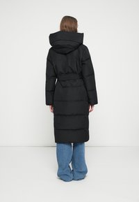 Žena zobrazená zezadu, oblečená v dlouhém černém prošívaném kabátu s kapucí a páskem a širokých modrých džínách na jednorodém bílém pozadí.