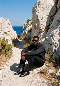 Άνδρας που φορά γυαλιά ηλίου και μαύρα ρούχα, καθισμένος σε πετρώδη μονοπάτι ανάμεσα σε μεγάλους λευκούς βράχους με τη θάλασσα στο φόντο.