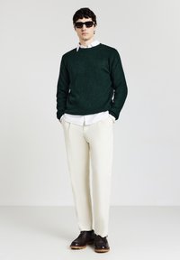 Uomo in maglione di filo verde scuro su camicia bianca, pantaloni crema, scarpe marroni scure e occhiali da sole neri in piedi contro uno sfondo bianco.