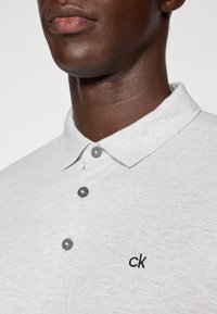 Camisa polo cinza em tecido texturizado, com um colarinho, três botões em cinza escuro e um subtil logótipo "ck" preto no peito.