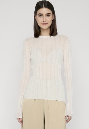 Kobieta o prostych ciemnych włosach nosi przezroczystą białą bluzkę z długim rękawem na białym staniku i beżowych spódnicach o szerokich nogawkach na jednolitym tle.