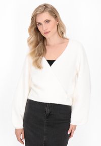 Femme blonde aux cheveux ondulés portant un pull enroulé blanc sur un top noir et une jupe noire taille haute, se tenant devant un fond blanc.