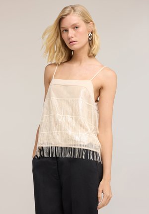 Femme blonde avec des boucles d'oreilles, portant un top sans manches beige à franges et un pantalon noir, debout devant un fond uni.