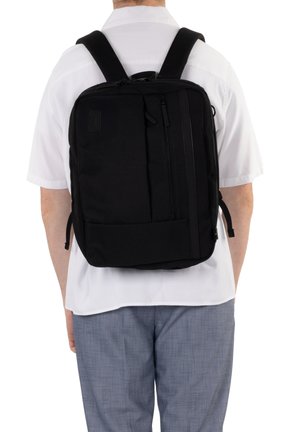 Homme portant un sac à dos noir rectangulaire avec plusieurs compartiments zippés, chemise blanche à manches courtes et pantalon gris, vu de dos.