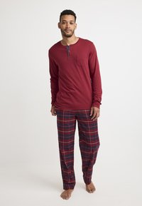Maglietta henley rossa a maniche lunghe con tasca, abbinata a pantaloni da pigiama scozzesi in rosso, blu e bianco. Materiale in cotone con una texture morbida.