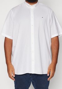 Uomo che indossa una camicia bianca a maniche corte, morbida, con un piccolo logo sul petto e pantaloni blu scuro.