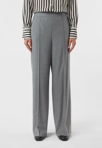 Kobieta w szarych, szerokich spodniach z wysokim stanem, do których dobrała długim rękawem koszulę w brązowo-białe pionowe paski oraz czarne buty.