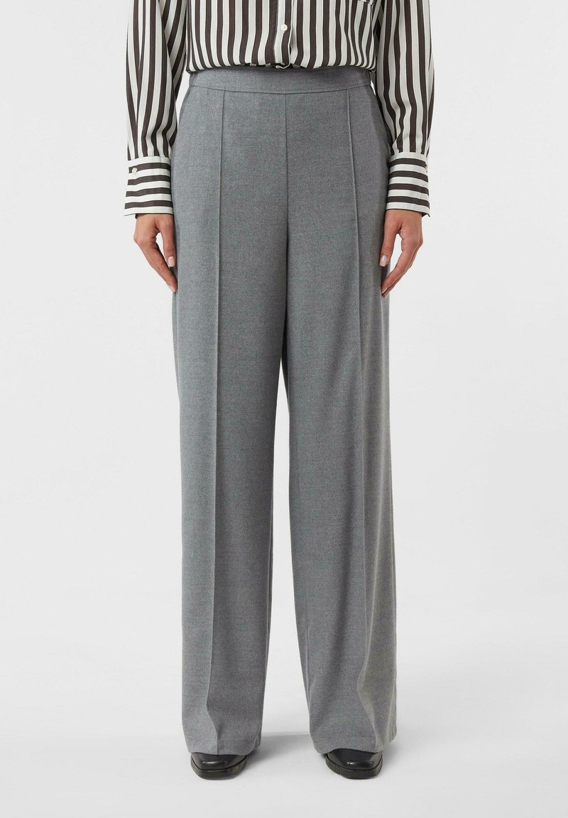 Kobieta w szarych, szerokich spodniach z wysokim stanem, do których dobrała długim rękawem koszulę w brązowo-białe pionowe paski oraz czarne buty.