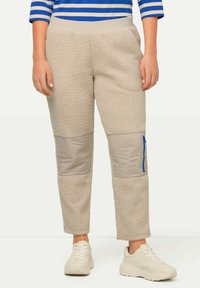 Beige Fleece-Hosen mit weicher Textur, ausgestattet mit beige farbenen Stoffpatches und einer blauen Reißverschlusstasche auf der rechten Seite, kombiniert mit hellen Sneakers.