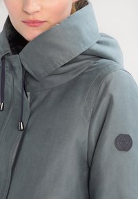 Grå hættejakke lavet af glat, vandafvisende stof. Har en høj krave, justerbare snore og en logo-patch på ærmet.