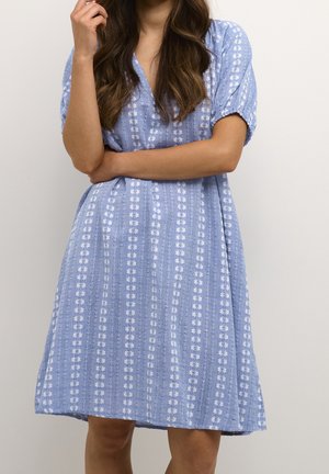 Femme portant une robe bleu clair à rayures verticales florales blanches, manches courtes et coupe ample, se tenant devant un fond clair uni.