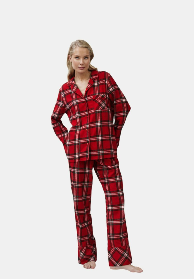 Vrouw die blote voeten staat en een rood en zwart geruit overhemd met lange mouwen en bijpassende losse broek draagt in een ontspannen houding.