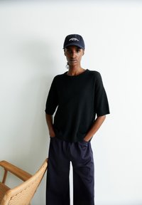 Schwarzer gerippter Strickpullover mit kurzen Ärmeln, kombiniert mit lockeren navyblauen Hosen. Eine navyblaue Mütze mit weißer Stickerei. Schlichte Kulisse und Stuhl.