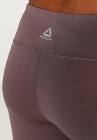 Šedé legíny s vysokým pasem, které mají hladkou texturu, jemný lesk a logo Reebok na pasu, navržené pro aktivní nošení.