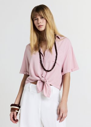 Camicia rosa a maniche corte annodata davanti, abbinata a pantaloni bianchi. Accessoriata con una collana di perline scure e braccialetti impilati.