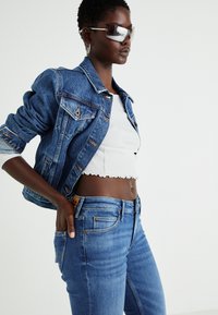 Model nosi modro denim jakno čez belo kratko majico in svetlo modre kavbojke. Obleka se ponaša s klasičnim dizajnom, ki ima priloženo silhueto.