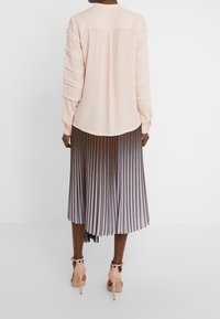Donna in piedi di spalle, che indossa una blusa rosa pallido con maniche a strati, una gonna midi plissettata metallizzata e sandali con tacco nudo.