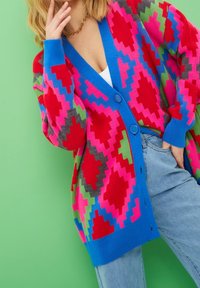 Kobieta w kolorowym, oversize'owym kardiganie z czerwonymi, różowymi, niebieskimi, zielonymi i czarnymi geometrycznymi wzorami, nosząca jasne dżinsy.