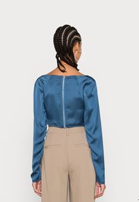 Top longo de cetim azul com decote quadrado e fecho exposto nas costas, combinado com calças bege de cintura alta.
