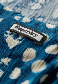 Tissu bleu à motifs avec des taches blanches, comportant une étiquette noir et blanc portant l'inscription "Superdry". Matériau en tricot texturé, vue en gros plan.