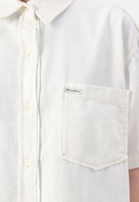 White denim short-sleeve shirt with a pointed collar, front buttons, and a single chest pocket featuring a Guess Jeans label.