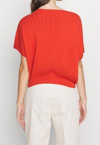 Top de punto rojo con canalé, de mangas anchas y cinturilla ajustada, combinado con pantalones de color claro, ligeramente texturizados.