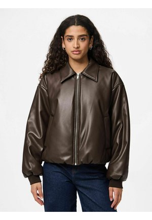 Femme aux longs cheveux bouclés portant une veste en cuir marron foncé zippée et un jean bleu, debout devant un fond blanc uni.