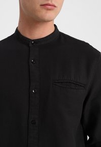 Svart skjorta med knappar och bandkrage, med en bröstficka, slät textur och subtil sömdetaljering.