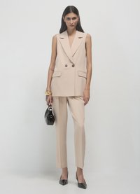 Beige Weste mit doppelten Knöpfen, kombiniert mit passenden taillierten Hosen. Eine schwarze Handtasche und spitze Absätze runden das Outfit ab. Schlichtes Hintergrund.