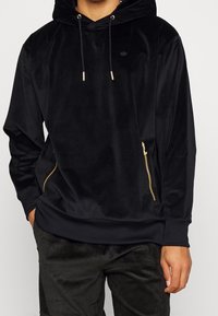 Svart sammetsluva med framficka och gyllene dragkedja. Har en huva med snörning och en liten logotyp på bröstet. Mjuk och slät textur.