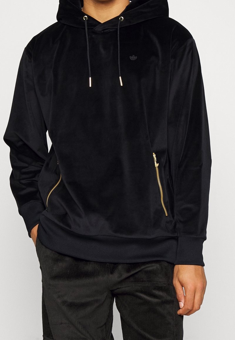 Svart sammetsluva med framficka och gyllene dragkedja. Har en huva med snörning och en liten logotyp på bröstet. Mjuk och slät textur.