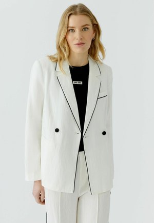 Vrouw met blond haar, gekleed in een wit pak met zwarte biezen over een zwart shirt met de tekst "OUI OUI" op een effen lichte achtergrond.