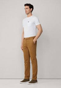 Camiseta blanca con bolsillo, chinos marrones y zapatillas verdes olivo. Los chinos cuentan con trabillas y una textura suave. Diseño sencillo.