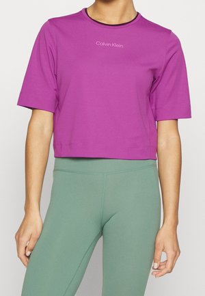 Woman wearing a purple cropped Calvin Klein T-shirt paired with high-waisted green leggings, standing against a plain background.