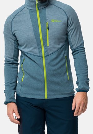 Mann trägt eine blaue Reißverschlussjacke mit limettengrünem Reißverschluss und Taschen, kombiniert mit dunkelblauer Hose, steht mit entspannten Armen.