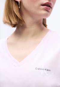 Γυναίκα με κοντά ξανθά μαλλιά που φοράει ανοιχτό ροζ μπλούζα Calvin Klein με V-λαιμόκοψη και χρυσό κρίκο σκουλαρίκι, κοιτάζοντας ελαφρώς προς τα δεξιά.