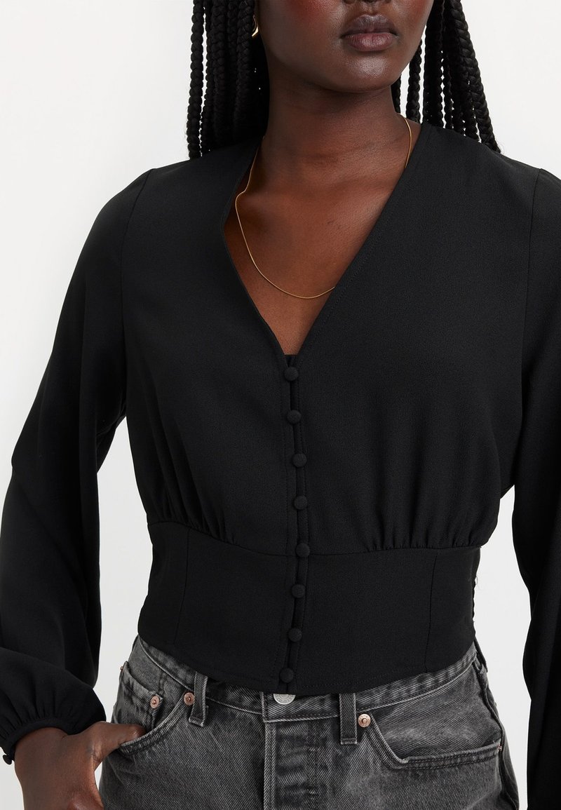 Schwarzes Langarmshirt mit V-Ausschnitt, aus strukturiertem Stoff, mit gerafftem Taillenbereich und Knopfdetaillierung vorne. Kombiniert mit grauen Jeans.